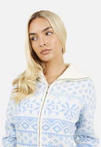 Lichtblauwe zip-up trui met een crèmekleurige kraag en ingewikkelde sneeuwvlok- en geometrische patronen in de stof gebreid.
