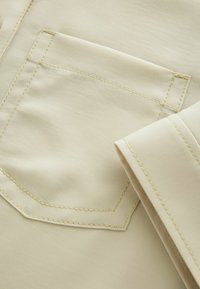 Tejido beige con una textura suave que presenta un bolsillo en el pecho y costuras amarillas en contraste. La prenda muestra líneas limpias y una manga doblada.