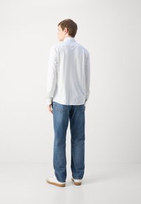 Camisa de manga longa branca com um padrão texturizado, combinada com jeans de ganga azuis e ténis brancos, vista de trás.