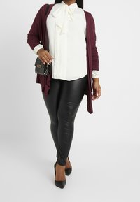 Femme portant une blouse blanche avec un nœud, un cardigan bourgogne, un legging en cuir noir, des talons noirs et portant un sac à bandoulière noir, se tenant sur un sol blanc.