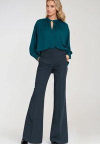 Kobieta stoi z dłońmi w kieszeniach, ubrana w turkusową bluzkę z długim rękawem i czarne szerokie spodnie z wysokim stanem na tle białym.