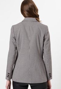 Blazer à carreaux noir et blanc, doté d'une coupe structurée, de revers en pointe et de poignets à trois boutons avec des accents marron. Texture lisse.