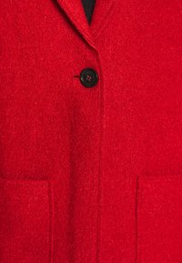 Manteau en laine rouge avec un bouton noir fermé à l'avant et deux poches carrées visibles près de l'ourlet inférieur.