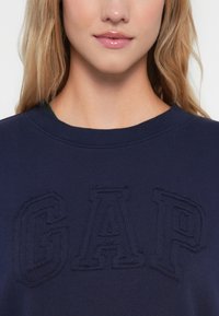 Tumepunasee navy sinine dressipluus ümmarguse kaelusega ja tikitud "GAP" logoga. Pehme kangas sujuva tekstuuriga ja lõdva istuvuse disain.