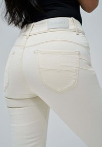 Jeans blancos ajustados con un diseño ceñido, que presentan un parche con el logo en la parte posterior de la cintura y dos bolsillos traseros con sutiles detalles de costura.