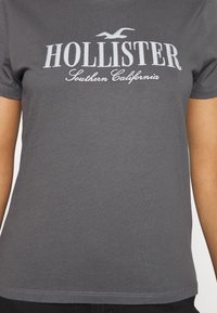 Szara bawełniana koszulka z krótkim rękawem i okrągłym dekoltem. Na przodzie znajduje się nadrukowany biały tekst "HOLLISTER" z napisem "Southern California" poniżej.