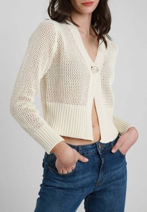 Donna con capelli scuri che indossa un cardigan crema lavorato a maglia aperta, abbottonato al petto, e jeans blu a vita alta con le mani nelle tasche.