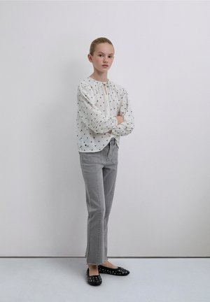 Blusa blanca de manga larga con lunares negros y lazo en el cuello; jeans grises de pierna recta; zapatos negros con tachuelas plateadas; fondo neutro.