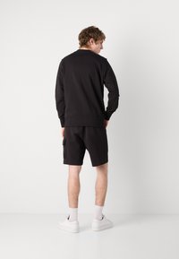 Felpa nera con polsini e orlo a costine, abbinata a pantaloni corti neri con tasche laterali. Il modello indossa sneakers bianche e calze.