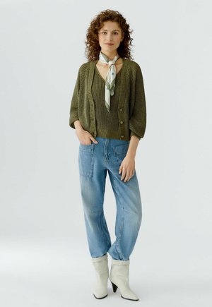 Vrouw met een olijfgroene gebreide cardigan en top, lichtblauwe losse jeans, witte laarzen met hak en een sjaal met patroon om haar nek, staand op een witte achtergrond.