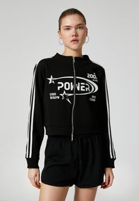 Sort kort sweatshirt med lynlås og hvide stjerner samt "POWER" logo. Har hvide striber langs ærmerne og er parret med sorte shorts.