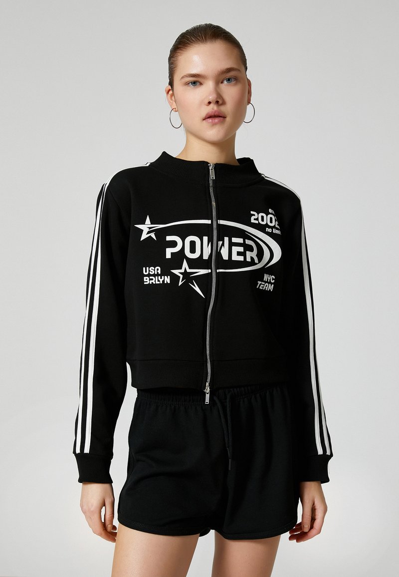 Sort kort sweatshirt med lynlås og hvide stjerner samt "POWER" logo. Har hvide striber langs ærmerne og er parret med sorte shorts.