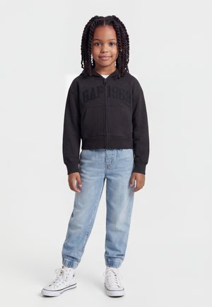 Mladé dievča s vrkočmi, oblečené v čiernej mikine na zips, svetlomodrých džínsoch a bielych vysokých teniskách, stojace proti bielemu pozadiu.