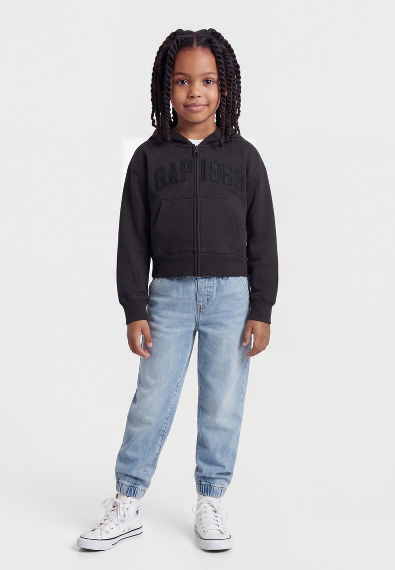 Jeune fille avec des tresses portant un sweat à capuche noir zippé, un jean bleu clair et des baskets blanches montantes, debout devant un fond blanc.