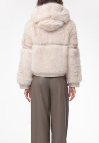Giacca in pelliccia sintetica di un leggero colore beige con cappuccio, caratterizzata da una consistenza morbida, polsini a coste e un design cropped. Indossata con pantaloni larghi marroni.