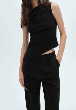 Kobieta ubrana w czarną, asymetryczną bluzkę bez rękawów i czarne spodnie, z jedną ręką w kieszeni, na jednolitym tle.