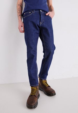 Mørkeblå denimjeans med kontrastsøm, med rett ben-design. Brukt med brune sko med snøring og gule lisseløsninger.