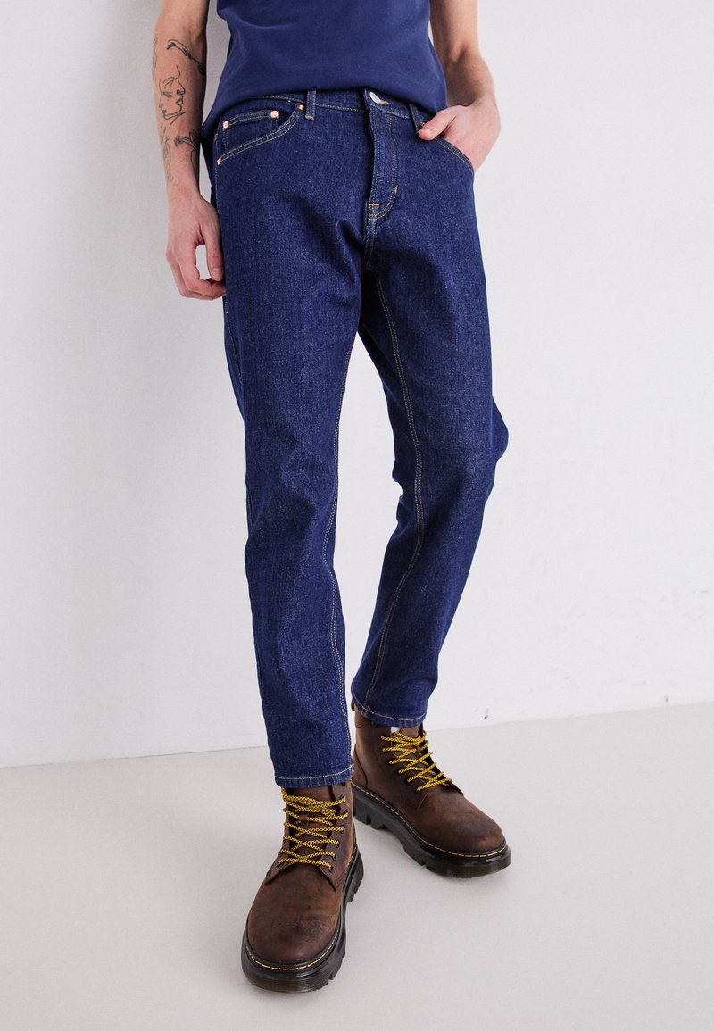 Mørkeblå denimjeans med kontrastsøm, med rett ben-design. Brukt med brune sko med snøring og gule lisseløsninger.