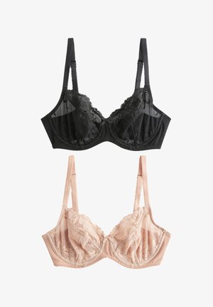 Smilende kvinde med langt brunt hår iført beige blonde-BH og matchende undertøj, står i et blødt oplyst rum med blomster og en sofa bag sig.