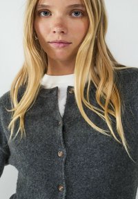 Graham Wolle cardigan mit Rundhalsausschnitt, ausgestattet mit fünf braunen Knöpfen und einer weichen Textur. Über einem weißen Hemd getragen.