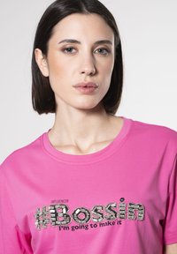 Camiseta rosa con cuello redondo y mangas cortas, con el atrevido texto en lentejuelas "#Bossin" y "INFLUENCER" impreso arriba.