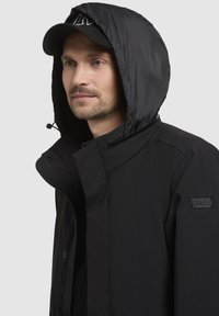 Chaqueta negra con capucha brillante y desmontable; cuenta con un cuello alto, cierre frontal con cremallera y parche con logotipo en la manga. Textura suave.