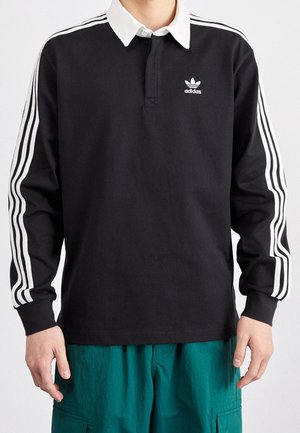 Osoba ubrana w czarną koszulkę polo Adidas z długim rękawem, białym kołnierzykiem i trzema białymi paskami na rękawach, w połączeniu z turkusowymi spodniami cargo.