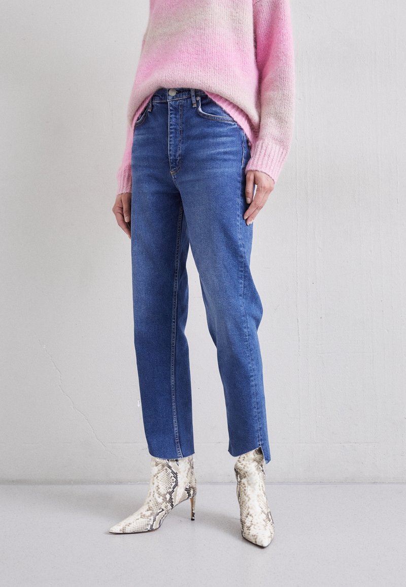 Femme portant un jean droit bleu, un pull ombré rose, et des bottes blanches à talons aiguilles en cuir de serpent.