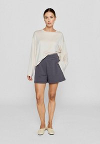 Hellbeige Langarmoberteil und dunkelgraue Shorts aus weichem Stoff, mit elastischem Bund und lockerer Passform. Hellfarbene flache Schuhe.