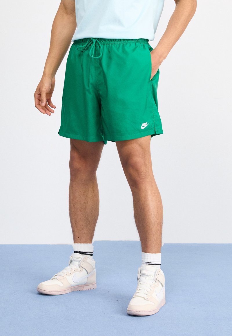 Personne portant un short vert Nike, des baskets blanches montantes et des chaussettes blanches, debout sur un sol bleu avec une main dans la poche.