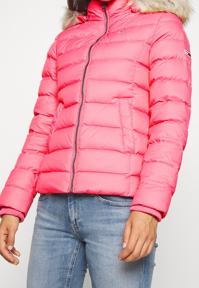 Polstrovaná růžová bunda s kapucí lemovanou kožešinou, má přední zip, boční kapsy a žebrované manžety. Hladký povrch, lehký design.