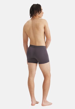 Giovane uomo con dreadlocks corti in piedi a piedi nudi, senza maglietta, indossa boxer grigio scuro, di spalle su uno sfondo bianco semplice.