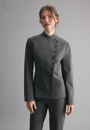 Femme portant un costume cintré gris foncé à fines rayures, avec un col montant et une fermeture asymétrique à boutons.