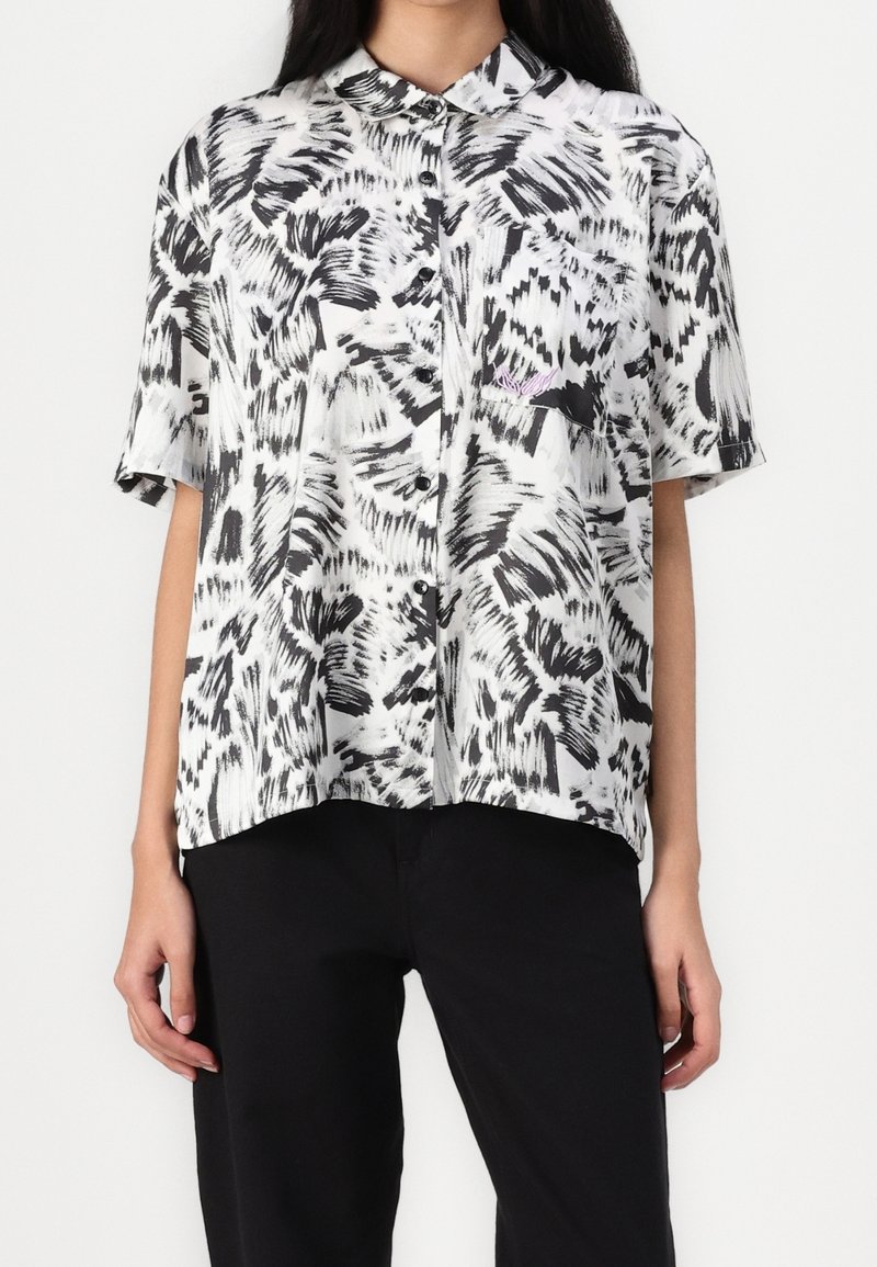 Chemise à manches courtes boutonnée avec un motif abstrait noir et blanc de coups de pinceau, portée avec un pantalon noir.