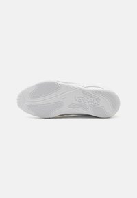 Suela de zapatilla blanca con goma texturizada, un intrincado patrón de relieve y el logo "VERSACE" grabado en el lateral.