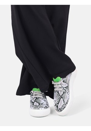 Person trägt schwarze Hosen mit weitem Bein und weiße Turnschuhe mit schwarzem Schlangenmuster und hellgrüner Zunge, steht mit überkreuzten Füßen.