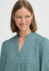 Svetlo teal blúzka s výstrihom do V, s plisovaným predným dielom a jemnými čiernymi vzormi mašlí. Materiál má krčenú textúru a zhromaždené detaily na pleciach.