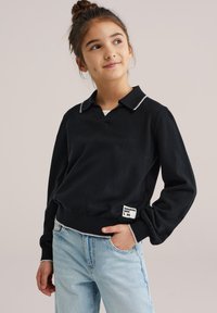 Jeune fille aux cheveux foncés attachés en chignon, portant un pull noir bordé de blanc et un jean bleu clair, souriante et regardant vers le haut à sa gauche.