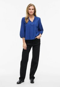 Blusa blu a maniche lunghe con scollo a V e dettagli con cordoncino, abbinata a pantaloni neri larghi e scarpe nere. Texture liscia.