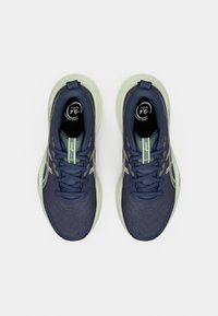 Pár sötétkék és világoszöld futócipő fűzőkkel, ASICS márkajelzéssel és címkével, amely a karbonlábnyom csökkentését jelzi.