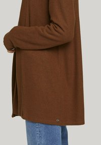 Brun långärmad topp i mjukt tyg med avslappnad passform, med en rundad nederkant och en liten logotypdetalj längst ner.