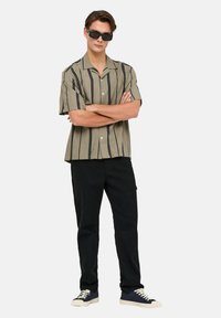 Short-sleeve, light olive green shirt with vertical black stripes, paired with black pants and navy sneakers. Model wearing black sunglasses.