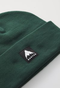 Zelená pletená čepice s ohnutým lemem, která má přišitou černou etiketu s grafikou hory a textem "BURTON". Měkká textura, jednoduchý design.