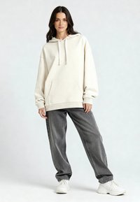 Noor naine seisab ühes ülekaalulises kreemjas kapuutsiga, lahtistes hallides teksades ja valgetes paksudes tossudes, lihtsa heleda tausta ees.