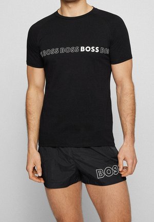 Mann trägt schwarzes T-Shirt mit wiederholtem "BOSS"-Text über der Brust und schwarze Shorts mit "BOSS"-Text am linken Oberschenkel.