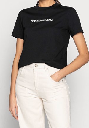 Henkilö pukeutuneena mustaan Calvin Klein Jeans -t-paitaan, joka on sullottu korkeavyötäröisiin luonnonvalkoisiin farkkuihin, toinen käsi taskussa, yksivärinen vaalea tausta.