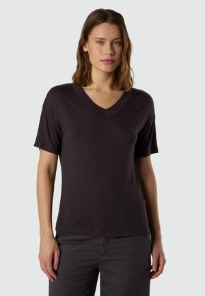 Jeune femme portant un t-shirt uni sombre à col en V et manches courtes, ainsi qu'un pantalon sombre, debout devant un fond gris clair.