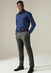 Camicia blu scuro a maniche lunghe; pantaloni grigi su misura; cintura e scarpe nere. Tessuto morbido, design aderente con dettagli a bottoni.