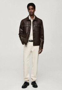 Veste en cuir brun avec col, deux poches poitrine et fermeture à boutons, associée à un pantalon clair et des chaussures noires.