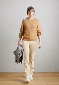 Burnt orange Wollsweater, creme Passformhose, weiße Sneakers; hält eine grau karierte Jacke. Glatte Texturen, lockere Passform, Grundfarben.
